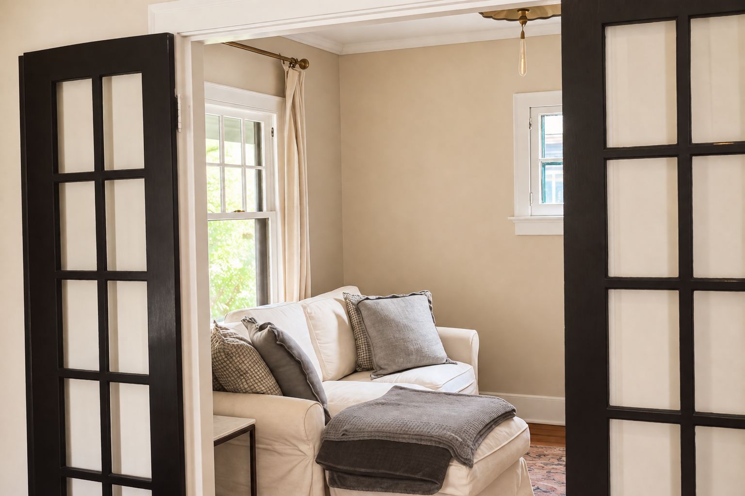 Una habitación con pared color crema, un sofá blanco con varios cojines, una mesita y una ventana con cortina.