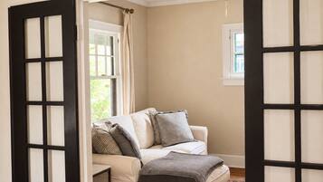 Una habitación con pared color crema, un sofá blanco con varios cojines, una mesita y una ventana con cortina.