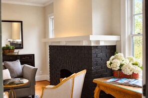 A living room with a fireplace, a sofa, a table with magazines, and a window with flowers. - St. Matthew’s - Walk to Shops & Restaurants (Louisville)