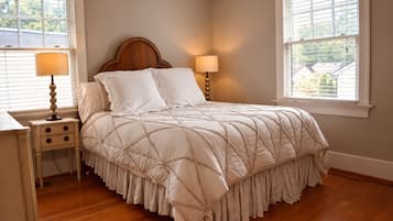 Een slaapkamer met een houten hoofdbord, een bed met een gewatteerde spreideken, twee bedlampjes, een nachtkastje met lades en een raam met jaloezieën.