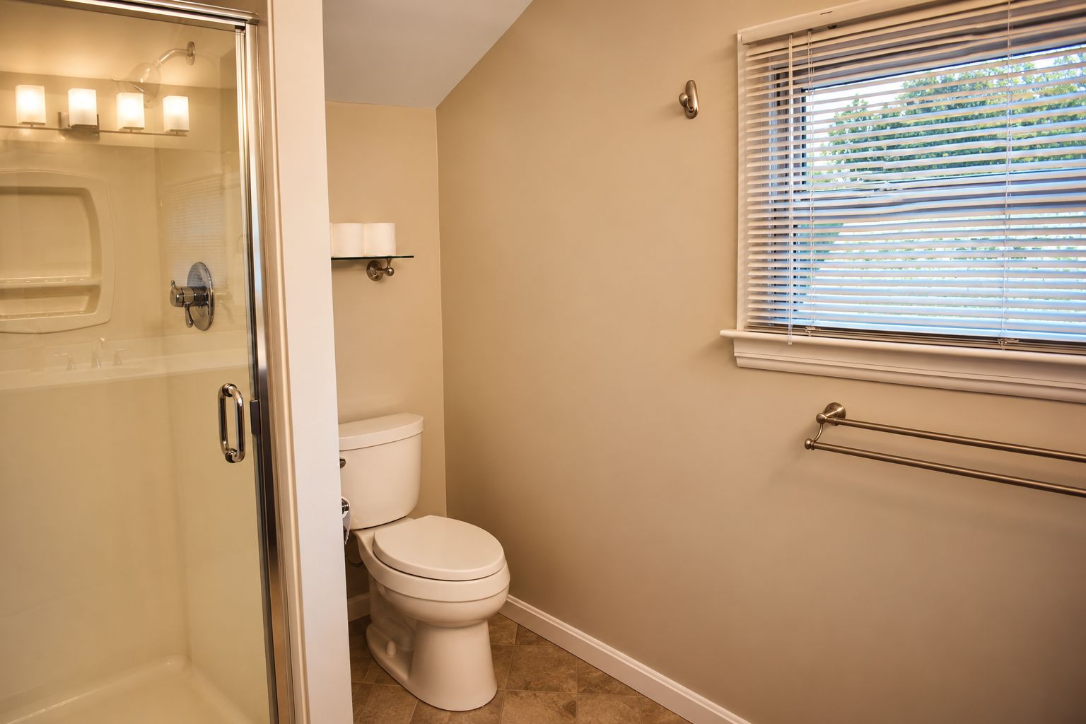 Ein Badezimmer mit Duschkabine aus Glas, Toilette, Handtuchhalter und Fenster mit Jalousien.