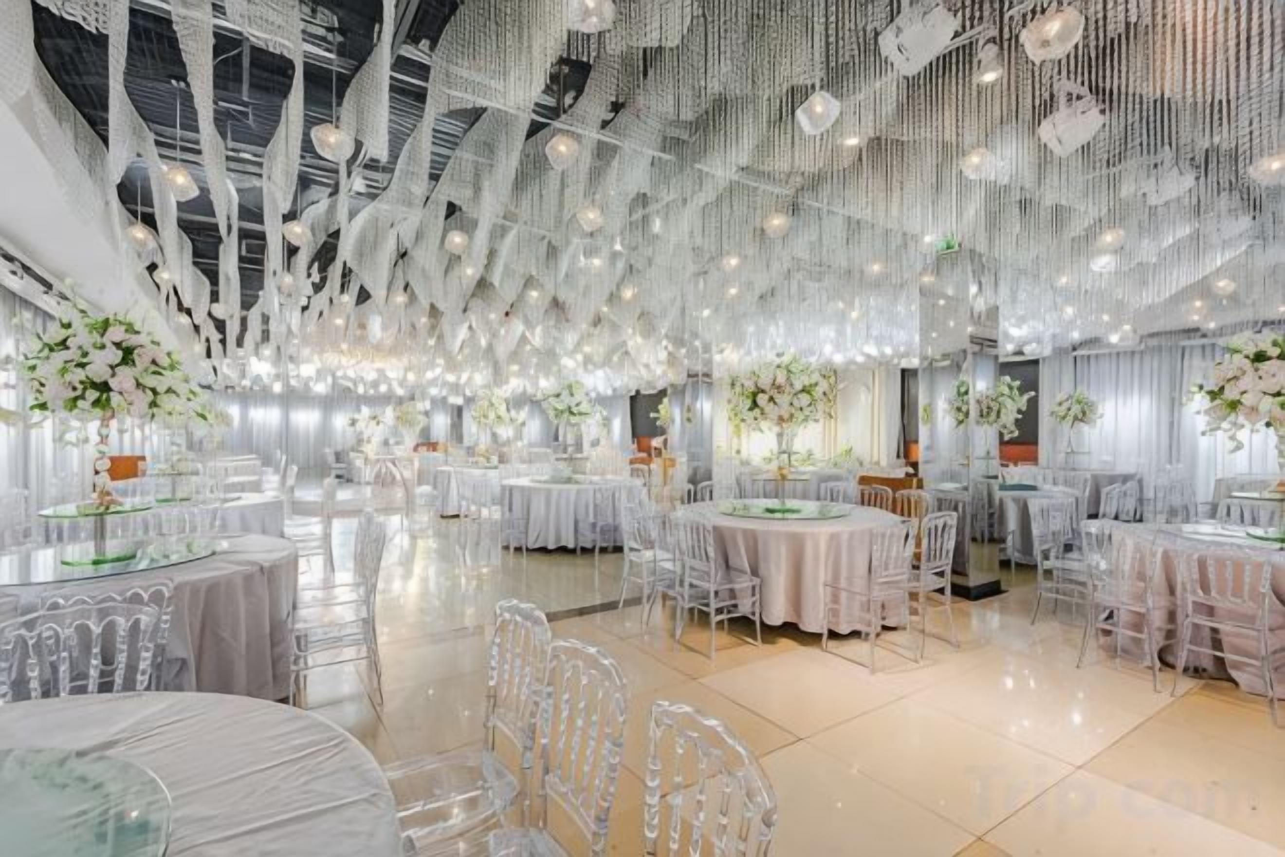 A spacious hall with multiple round tables, clear chairs, and hanging light fixtures.