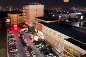 Ein mehrstöckiges Hotel mit Parkplatz voller Autos, einer großen beleuchteten Schautafel und einer Stadtansicht im Hintergrund.