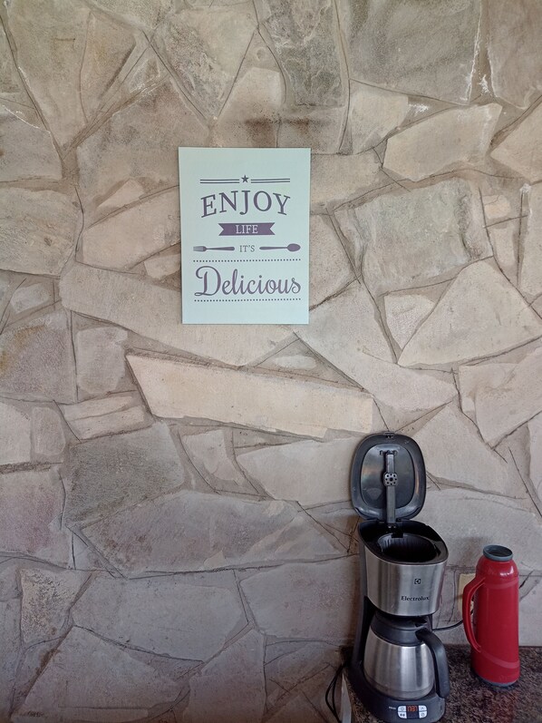 Un muro de piedra con un letrero que dice 'ENJOY LIFE IT'S Delicious' y una cafetera con un termo rojo a su lado.
