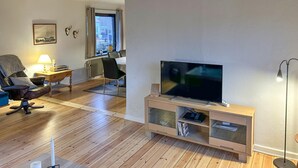 A living room with a wooden floor, a TV on a glass cabinet, a chair, a lamp, and a window with curtains. - Beautiful home in Aabenraa with WiFi (Aabenraa)
