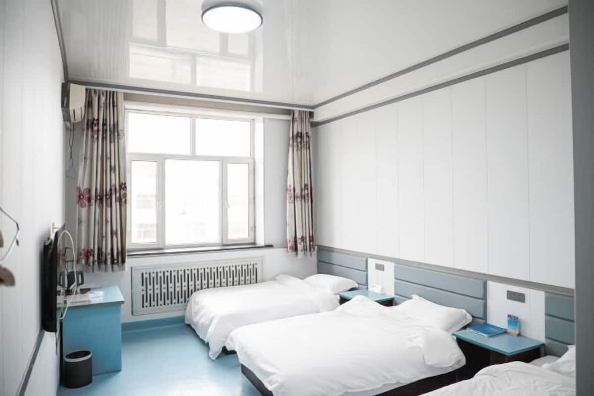 A hotel room with two beds, a window with curtains, a radiator, a desk with a chair, and a nightstand with a lamp.