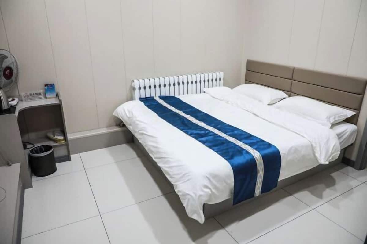 A hotel room with a bed, a radiator, a small table with a fan, and a trash bin.