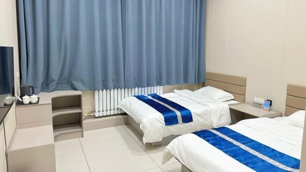 A hotel room with two beds, a desk, a chair, a radiator, and a window with curtains. - Huma Hongyue Hotel (Huma)