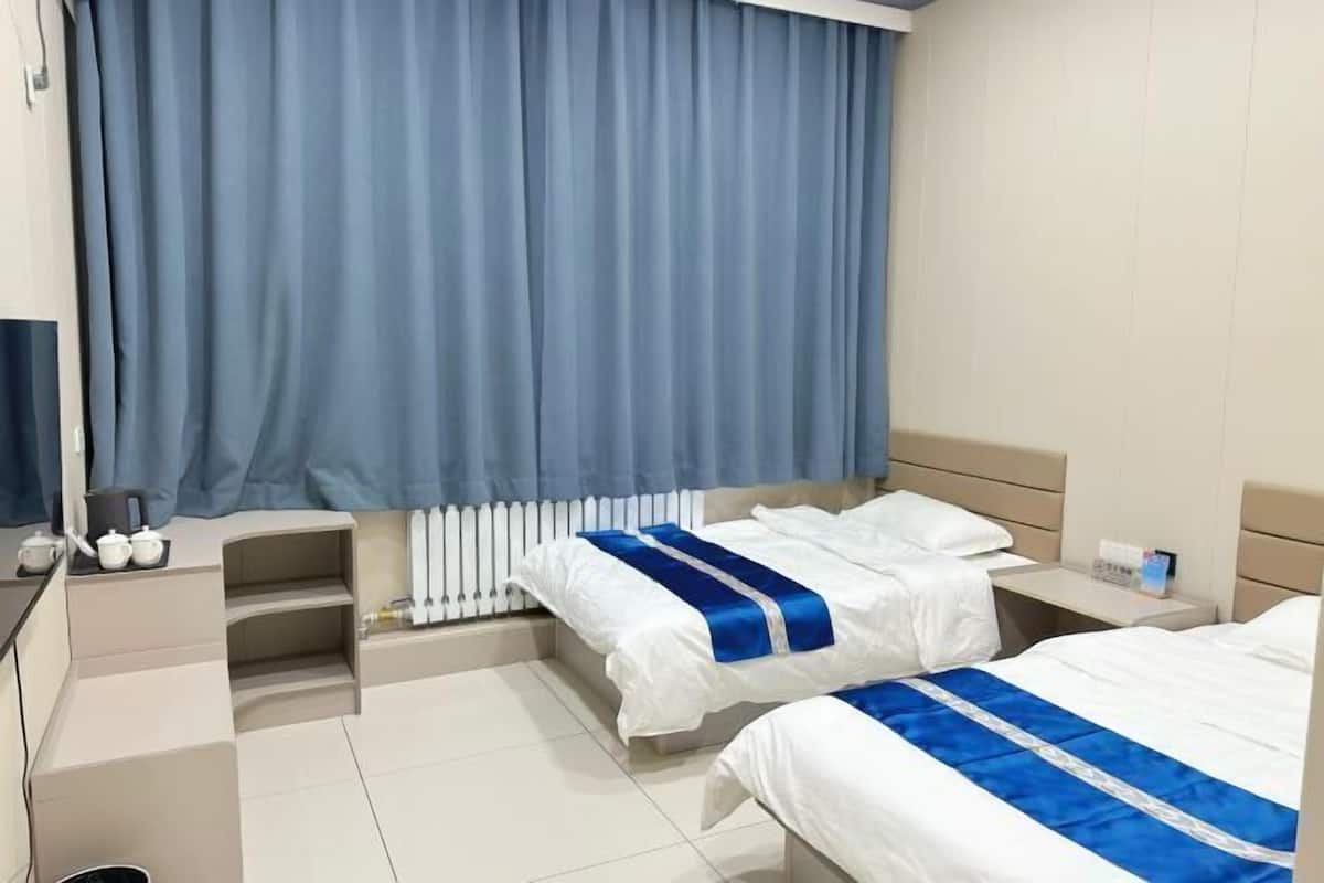 A hotel room with two beds, a desk, a chair, a radiator, and a window with curtains.