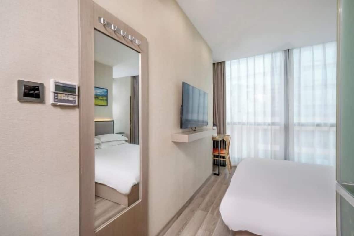 Een hotelkamer met een bed, een aan de muur bevestigde televisie, een bureau met stoel en een groot raam met gordijnen.