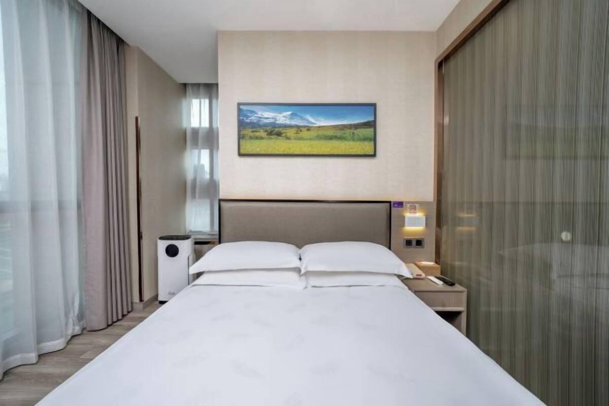 Een hotelkamer met een bed, nachtkastje, lamp, schilderij en een raam met gordijnen.