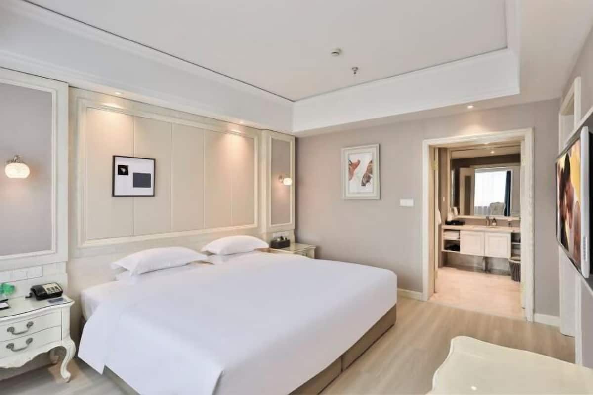 Une chambre d’hôtel avec un grand lit, des tables de chevet, une armoire, un miroir et une salle de bain visible à travers une porte ouverte.