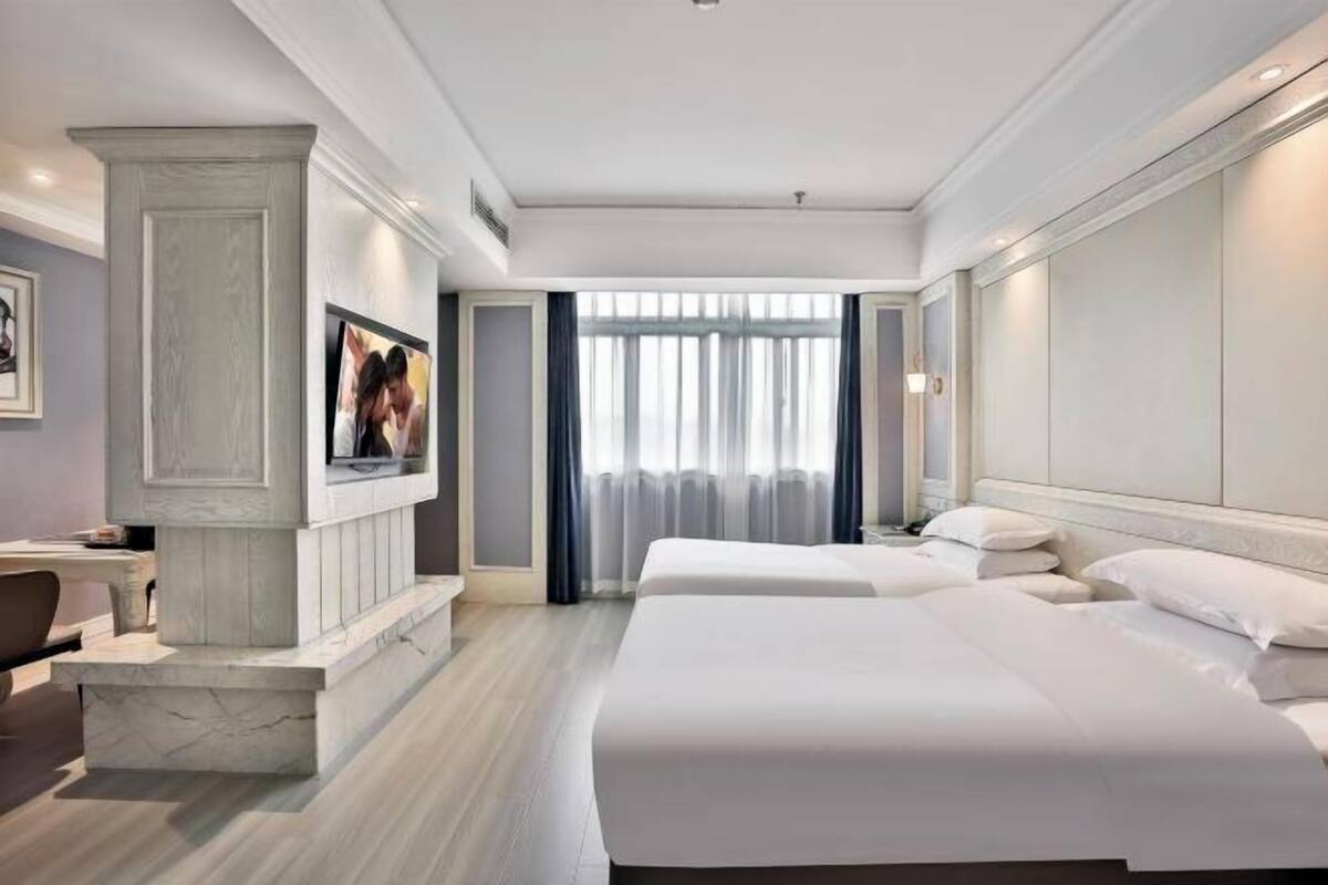 Une chambre d’hôtel avec deux lits, un bureau, une chaise, une grande fenêtre avec des rideaux et un tableau encadré au mur.