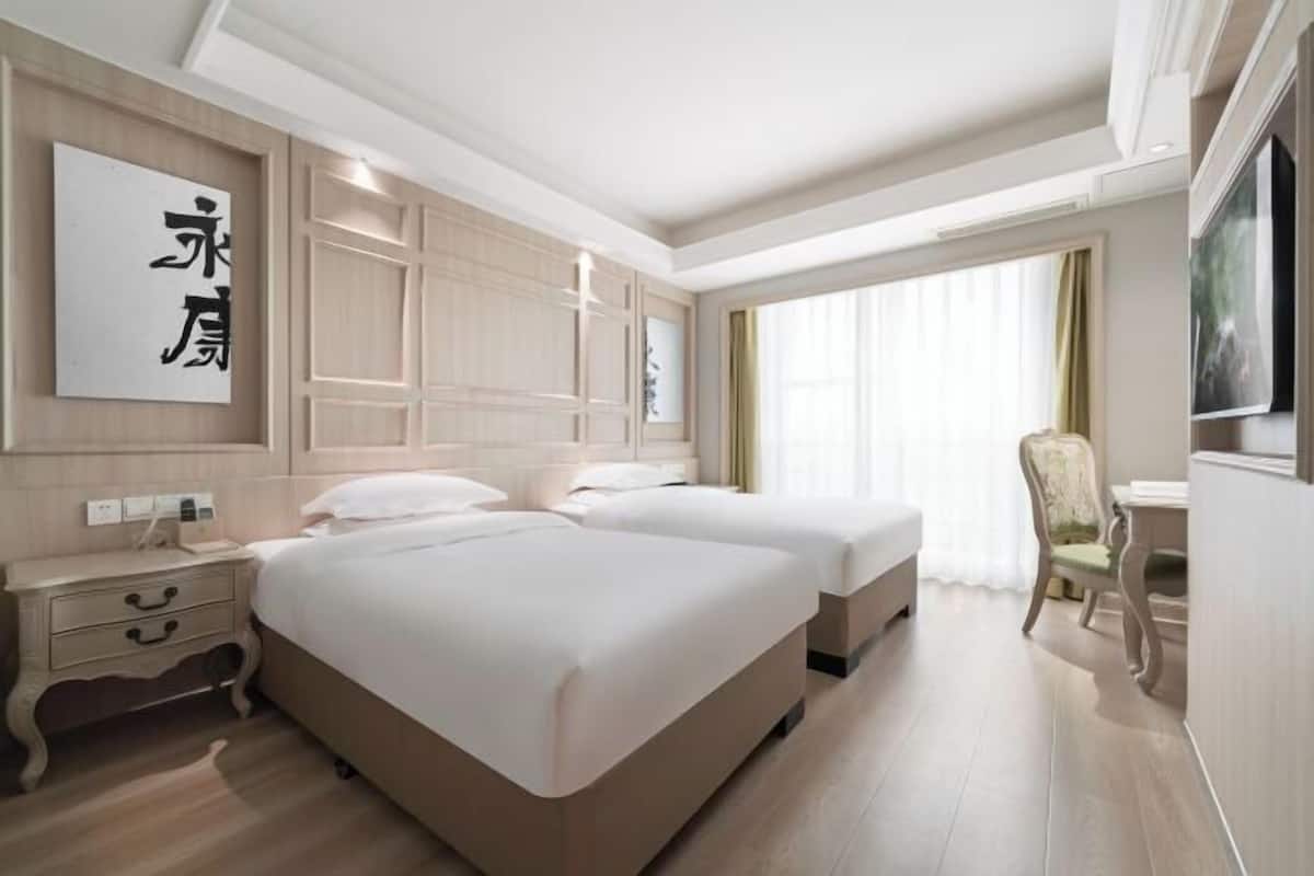 Une chambre d’hôtel avec deux lits, un bureau, une chaise et une grande fenêtre avec des rideaux.