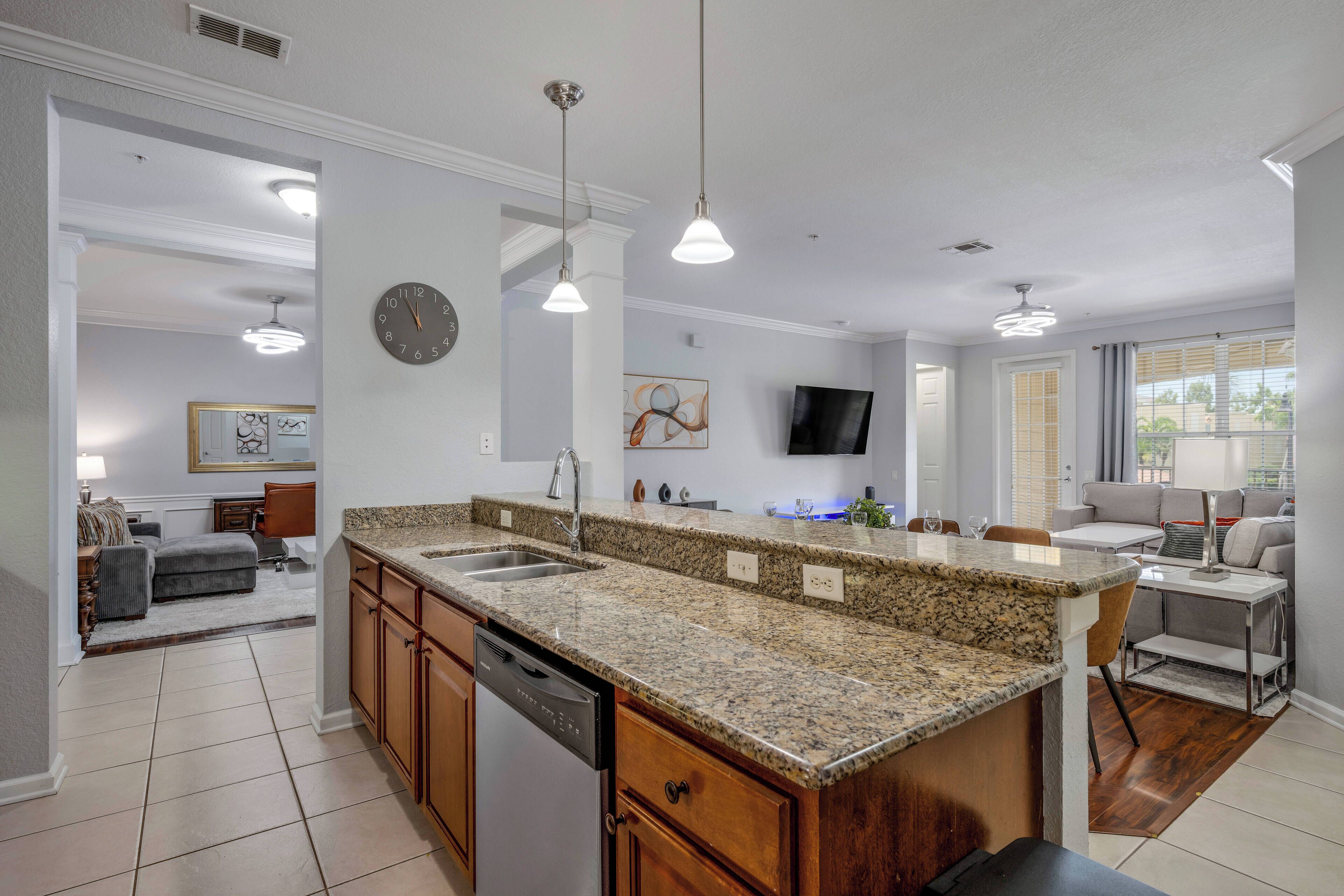 A modern kitchen with granite countertops, a dishwasher, and a dining area with a gray sofa and a TV mounted on the wall.