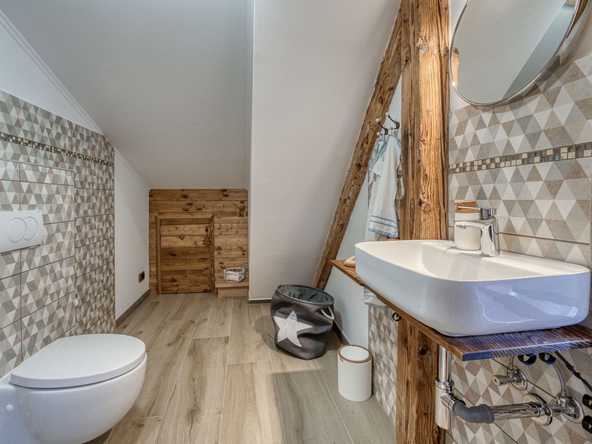 A bathroom with a wooden floor, a white toilet, a sink with a mirror, and patterned tiles on the walls.