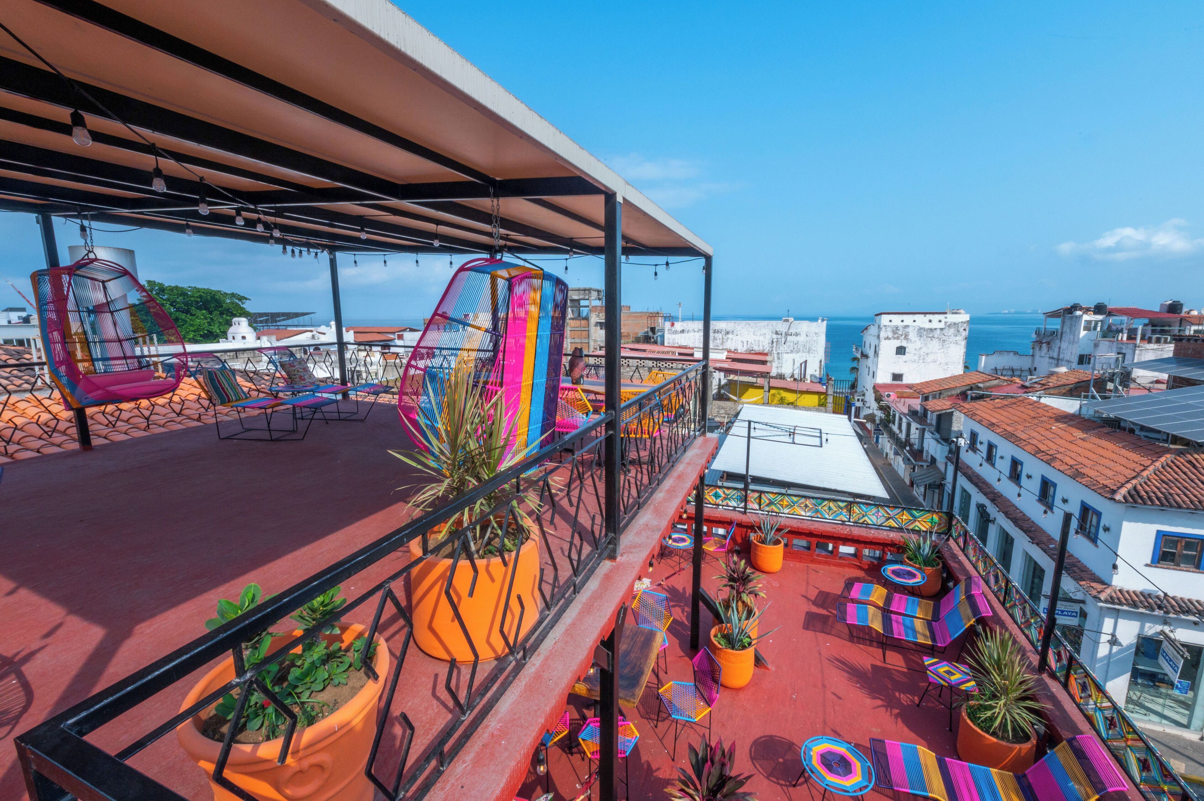 A rooftop terrace with colorful seating, potted plants, and a view of the city and sea.