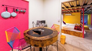 Ein Zimmer mit einem runden Holztisch, bunten Stühlen, einem Bett mit Holz Kopfteil und einer Küchenzeile mit hängenden Töpfen.
