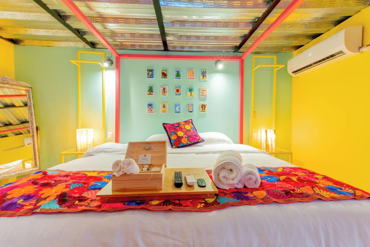 Une chambre d’hôtel avec un lit, un plateau contenant des serviettes et des télécommandes, un mur décoré d’œuvres d’art colorées et un appareil de climatisation.