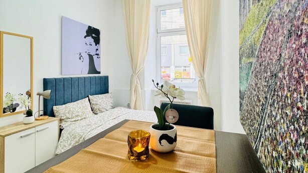 Un dormitorio con cama, una mesita de noche con lámpara, una ventana con cortinas, una pared con un cuadro grande, y vista a la calle.
