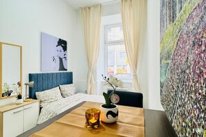 Ein Schlafzimmer mit Bett, Nachttisch mit Lampe, Fenster mit Vorhängen, einer Wand mit einem großen Gemälde und Blick auf die Straße.