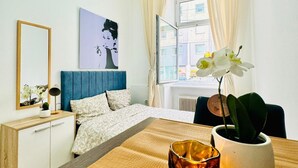 Un dormitorio con cama, mesita de noche, espejo, ventana con cortinas y una planta en maceta.
