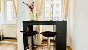 Une pièce avec une fenêtre, un radiateur, une table avec deux tabourets et un vase de fleurs.
