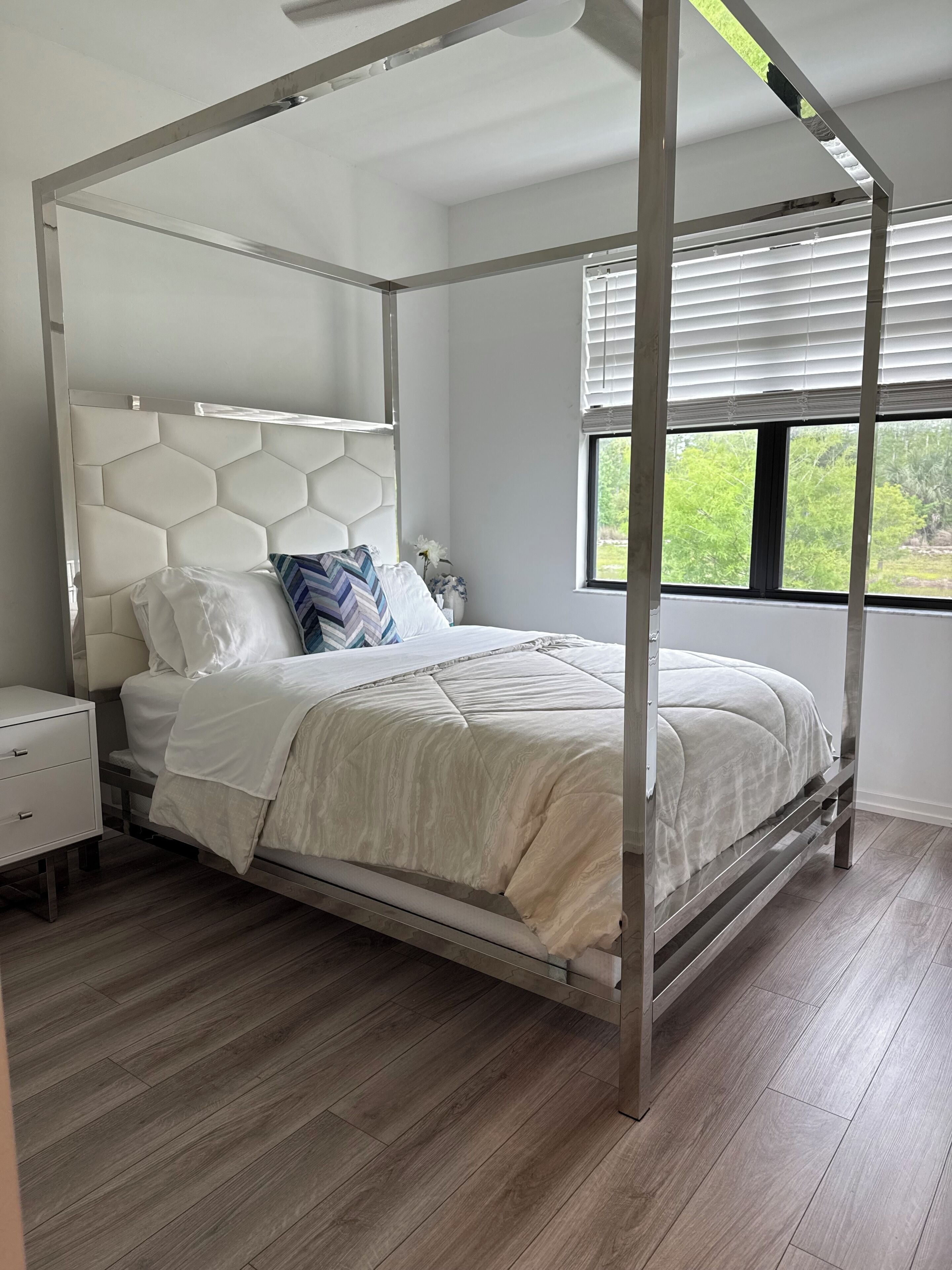 A four-poster bed with a hexagonal headboard, a bedside table, and a window with blinds.