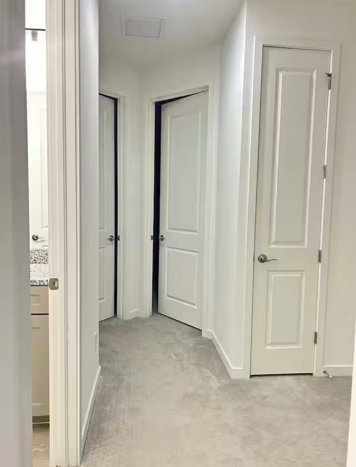 A narrow hallway with white doors, carpeted floor, and a visible kitchen area with granite countertops.