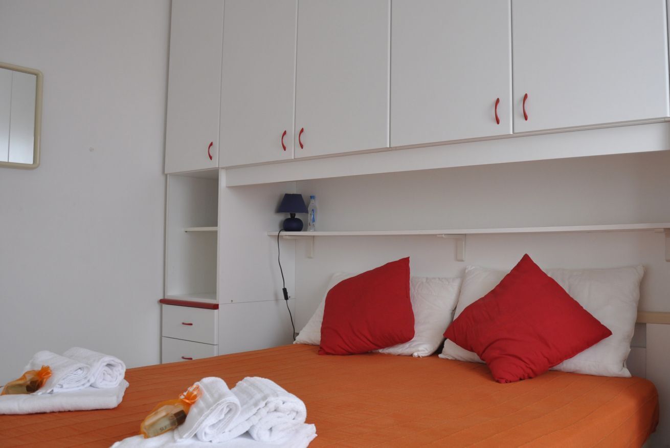 Una habitación de hotel con cama, almohadones rojos, un armario blanco y una mesita de noche con lámpara.