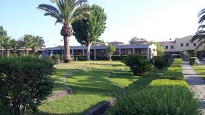 A landscaped garden with palm trees and neatly trimmed bushes, a paved walkway, and a building with a flat roof in the background. - La Conchiglia  - Piscina e Animazione gratuita (Bernalda)