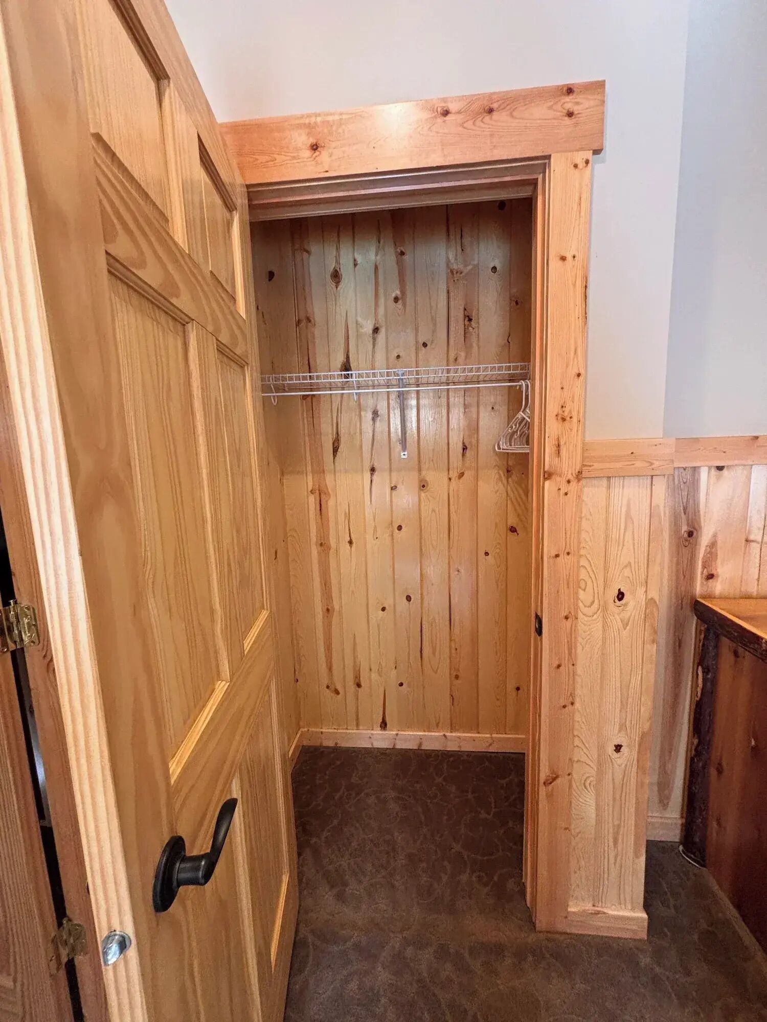A wooden closet with a metal rod shelf, a door with a black handle, and a carpeted floor.