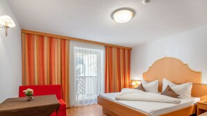 Ein Hotelzimmer mit einem hölzernen Bett, einem roten Sessel, einem kleinen Tisch mit einer Vase und einem Fenster mit Vorhängen.