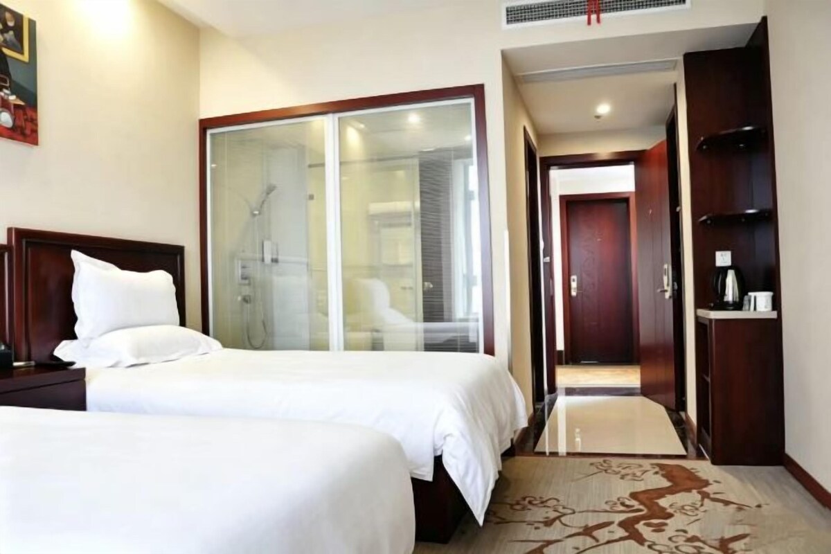 A hotel room with two beds, a glass shower enclosure, a wooden wardrobe, and a carpeted floor.