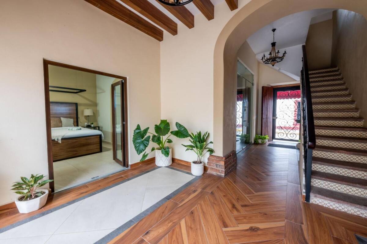 A hotel hallway with a wooden floor, a bed with a headboard, potted plants, and a staircase with a patterned railing.