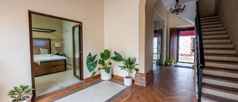 Pasillo de hotel con piso de madera, cama con cabecera, plantas en macetas y una escalera con barandilla decorada.