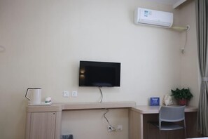 Et hotelværelse med et fladskærms-tv monteret på væggen, et skrivebord med stol, en lille potteplante og en aircondition-enhed.