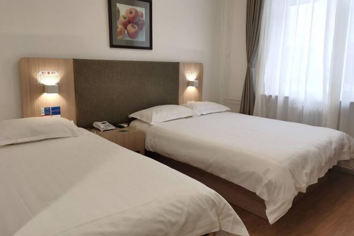 A hotel room with two beds, a wooden headboard, bedside lamps, a framed picture on the wall, and a window with curtains.