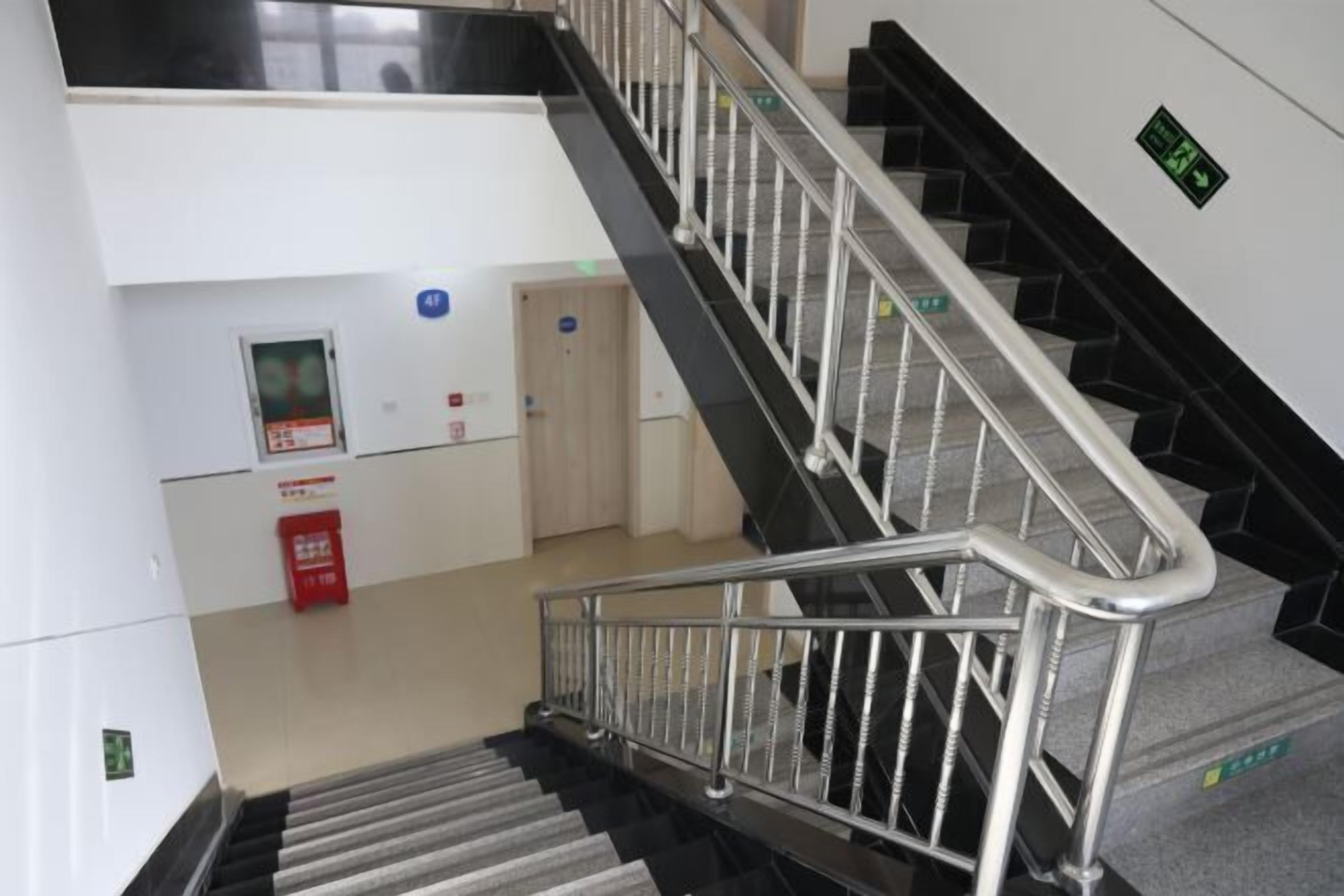 A staircase with metal railings leading to an upper floor. There is a fire alarm panel on the wall to the left and a trash bin below the stairs.