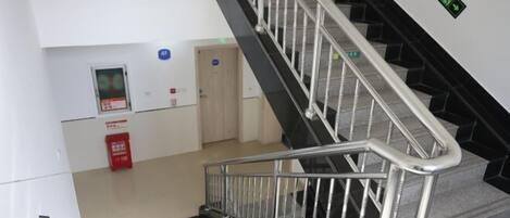 A staircase with metal railings leading to an upper floor. There is a fire alarm panel on the wall to the left and a trash bin below the stairs.