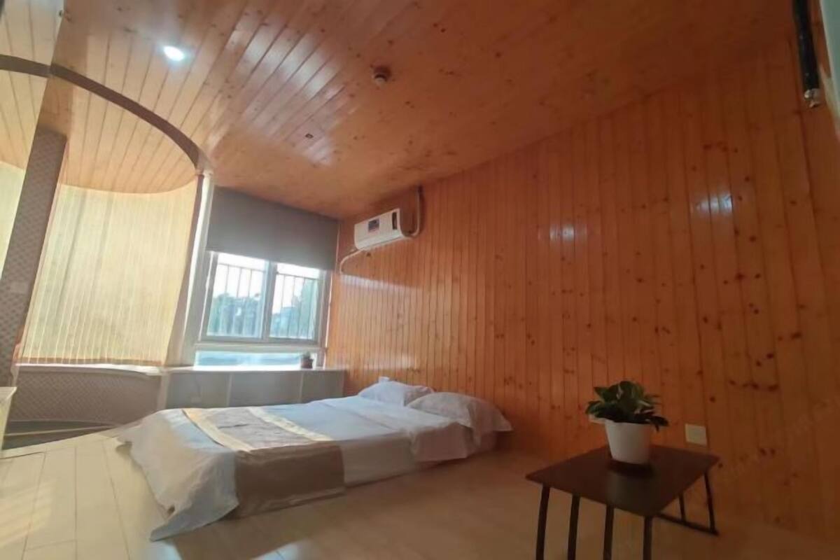 Une pièce aux murs en bois, un lit avec une literie blanche, une petite table avec une plante et une fenêtre avec des stores.
