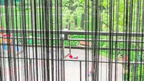 Une vue à travers une fenêtre munie de stores verticaux, dévoilant un balcon avec une balustrade et un espace extérieur verdoyant, agrémenté d’arbres et d’un banc.