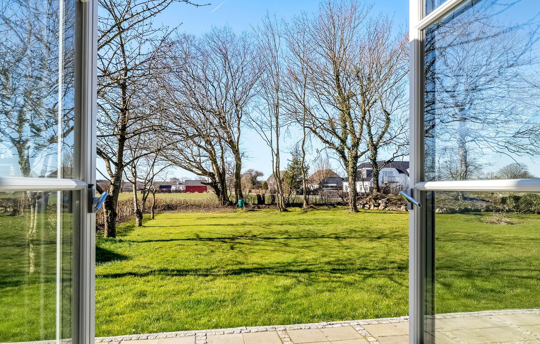 Uitzicht door een raam op een gazon met bomen en een rood gebouw op de achtergrond.