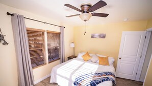 Et soveværelse med seng, ventilator, et vindue med persienner, en dør og et natbord med lampe.