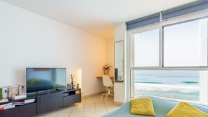 Moderni hotellihuone, jossa on suuri ikkuna, josta avautuu näkymä merelle, sinisillä ja keltaisilla tyynyillä varustettu sänky, työpöytä tuolilla sekä tummalla alustalla oleva televisio.