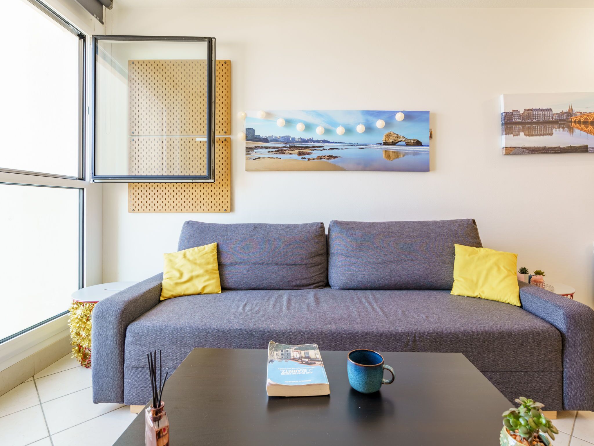 A modern living room with a grey sofa, yellow cushions, a black coffee table, a blue mug, and a framed cityscape painting.