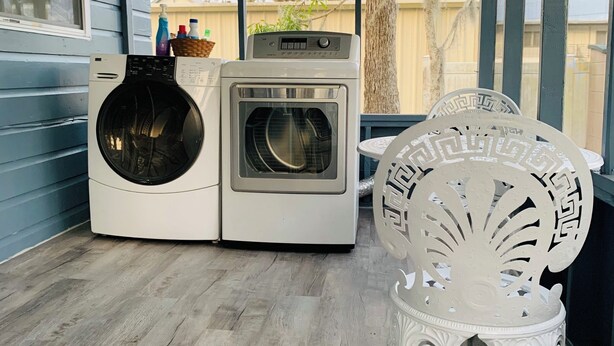 A laundry room with a front-loading washer and dryer, a decorative white bench, and large windows offering a view of the outdoors. - The Studio on 8th, Entire guest suite in Wauchula Florida ! (Wauchula)
