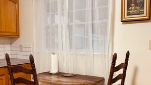 A dining area with a round table, two wooden chairs, a white curtain, a framed picture on the wall, and wooden cabinets. - The Studio on 8th, Entire guest suite in Wauchula Florida ! (Wauchula)