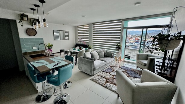 Eine moderne Küche mit Essbereich, ein graues Sofa und Blick auf die Stadt durch große Fenster.