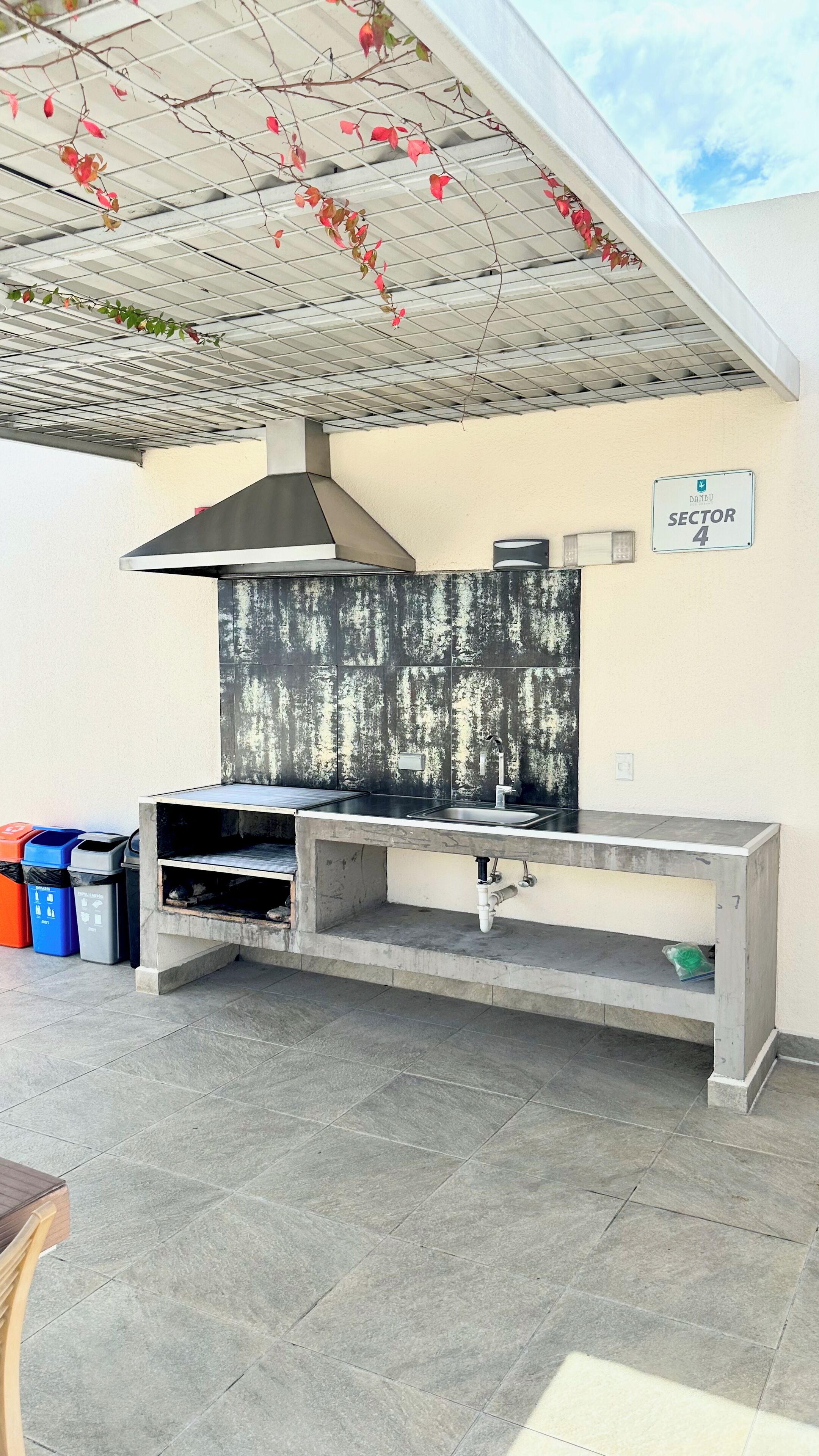 Un espace cuisine extérieur doté d’un évier en acier inoxydable, d’un pare-feu en carreaux noirs et d’une hotte aspirante. On y trouve également des poubelles et un panneau indiquant « Secteur 4 ».